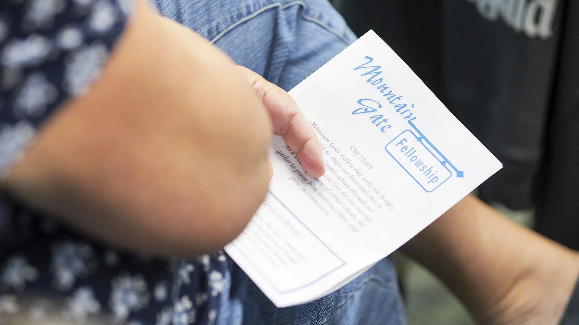 Person holding a flyer titled 'Mountain Gate Fellowship Our Vision' with blue and black text, wearing a blue patterned shirt and jeans, sitting in a location with blurred background.