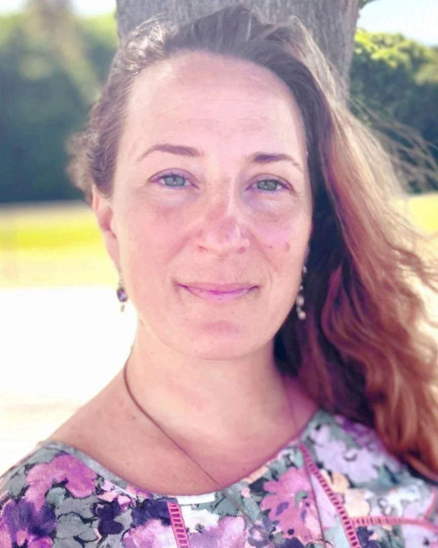 A woman with long, wavy red hair, blue eyes, and light skin, smiling outdoors near a tree in bright sunlight. She is wearing earrings, a floral top, and a black necklace.