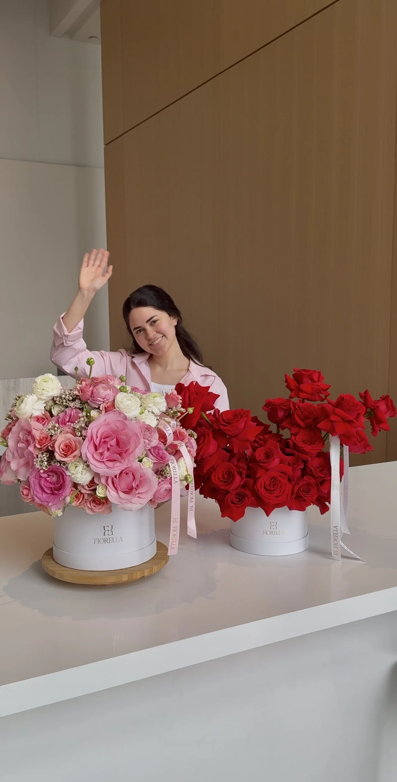 Smile girl in pink shirt smiling and waving, sitting behind two white boxes of flowers with pink and red roses, on a white table in a modern room.