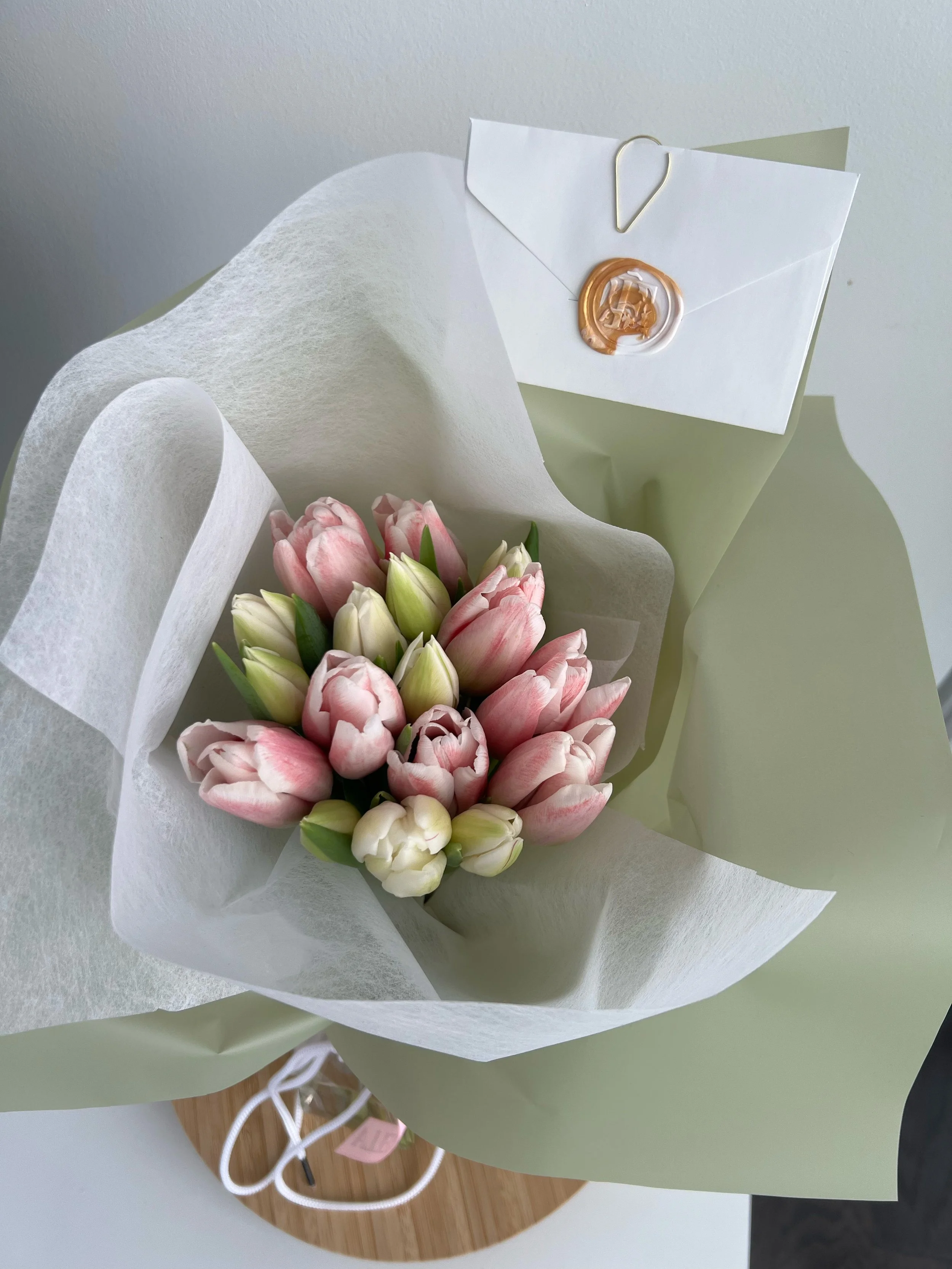 A bouquet of pink and white tulips wrapped in white and light green paper, with a white envelope with a gold wax seal and a small paperclip attached to it.
