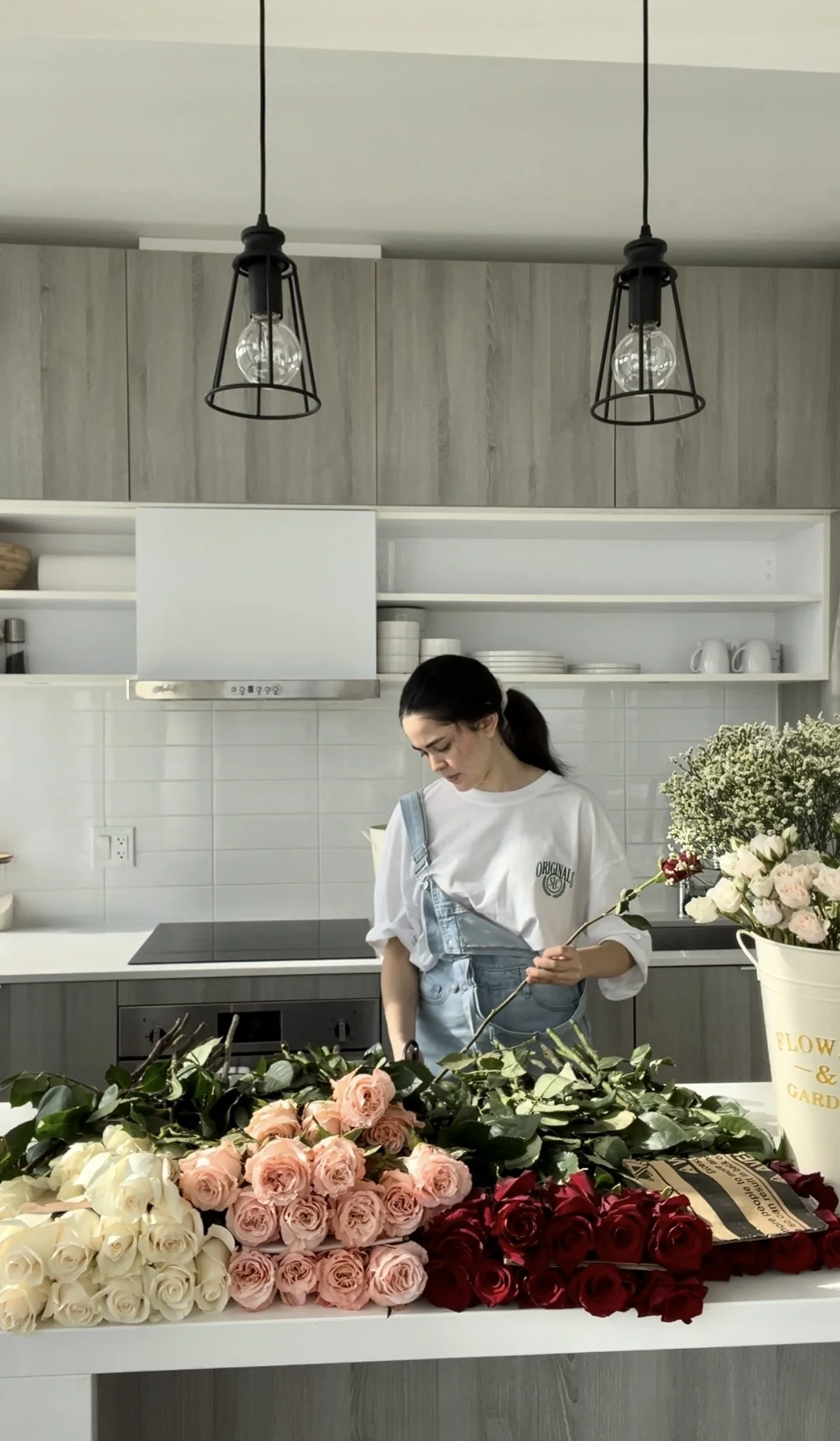 A woman preparing floral arrangements in a modern kitchen with a table of pink, white, and red roses in the foreground.