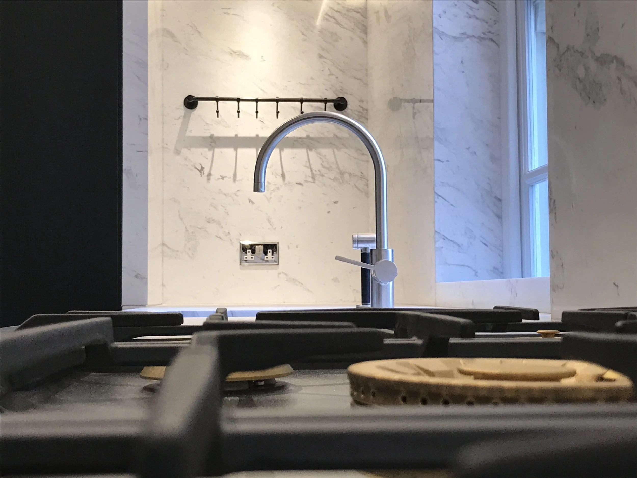 View of a modern kitchen sink with a chrome faucet in front of a light marble backsplash wall and a window to the right. The foreground shows the back of dark chairs and a table.