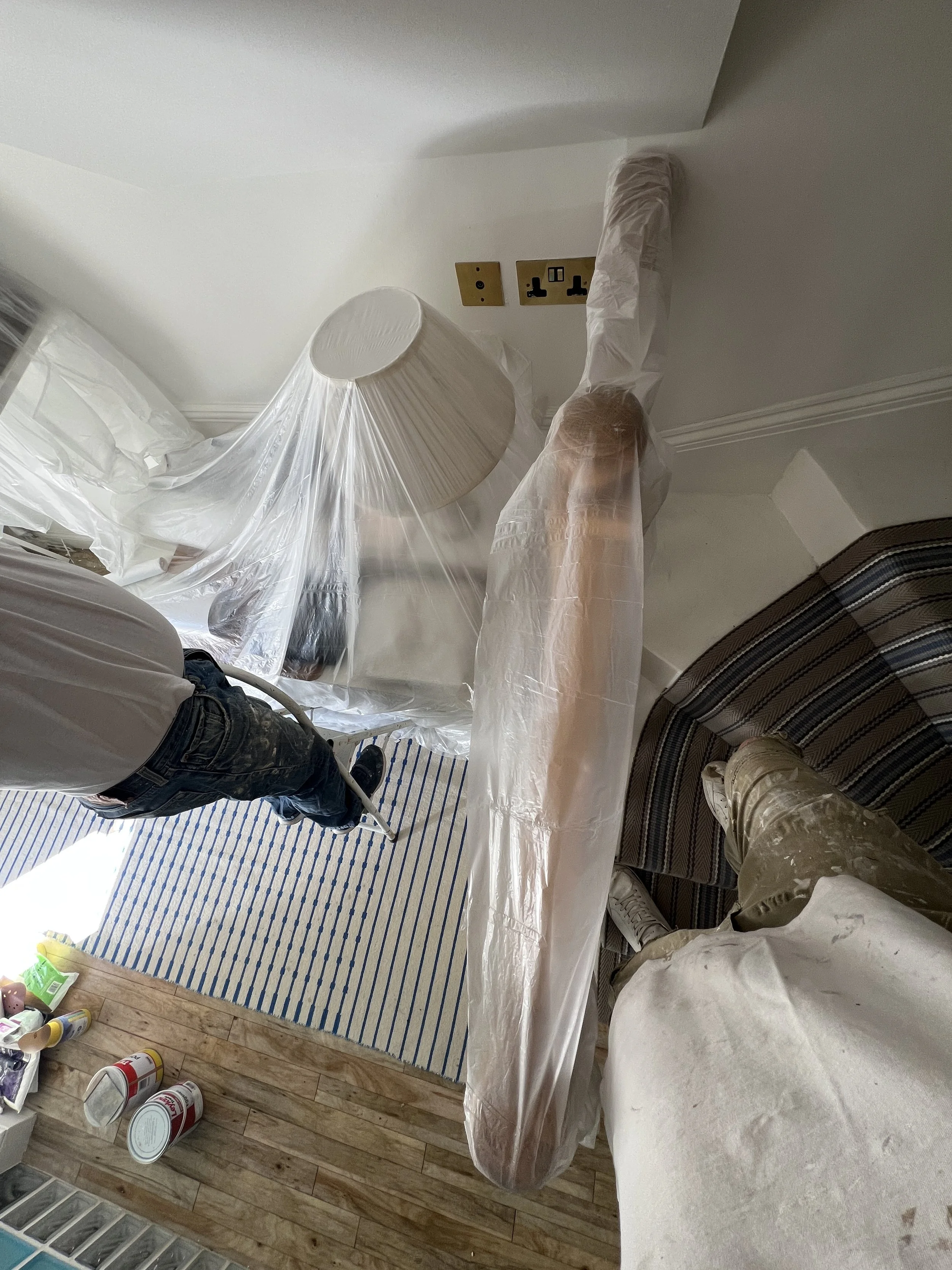 Image shows a room with a man and a woman preparing painting or renovation work. Furniture and supplies are covered with plastic. The floor has a striped rug, and there are paint cans on the floor.