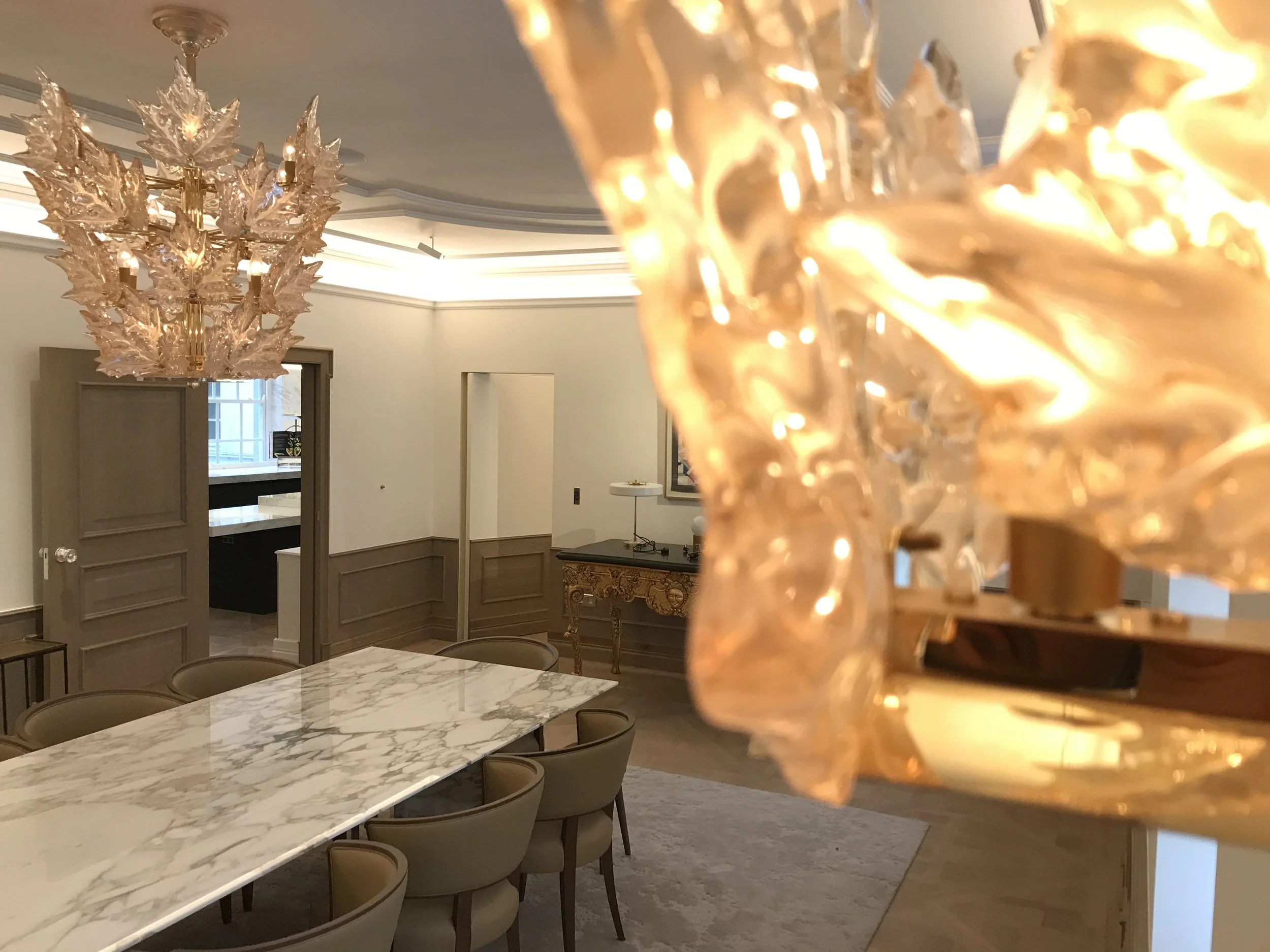 Elegant dining room with a marble-top dining table and beige chairs, a gold and glass leaf chandelier hanging from the ceiling, and a decorative table against the wall.