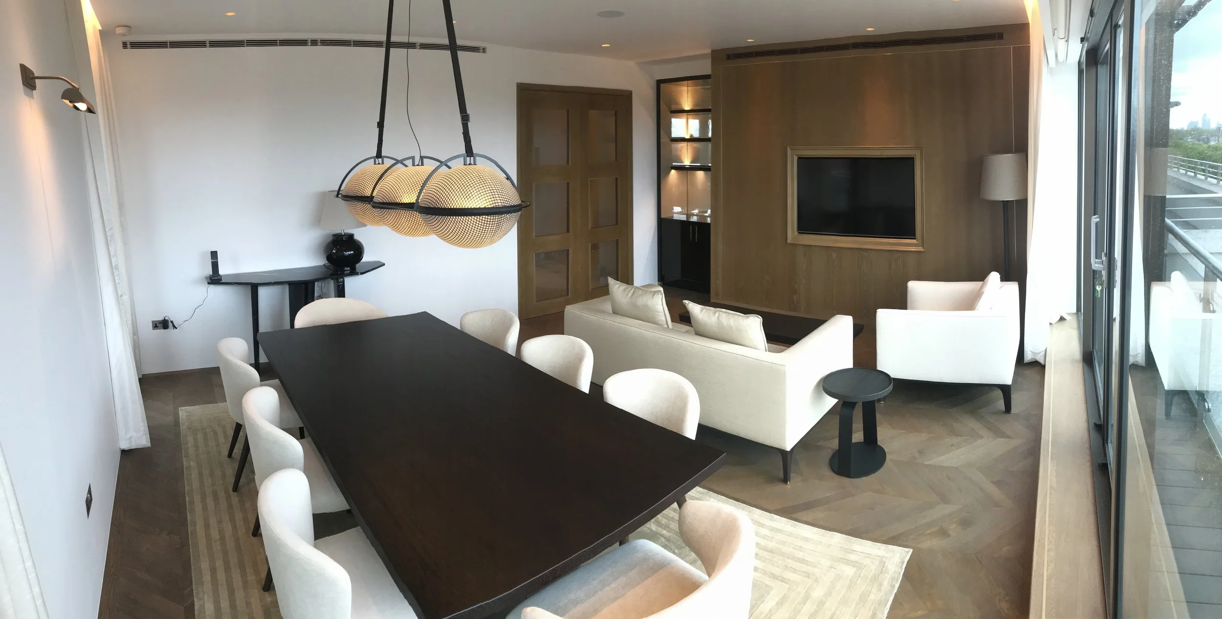 Modern living and dining room with white sofas, black dining table with white chairs, large windows, wall-mounted TV, wooden accents, and a decorative light fixture.