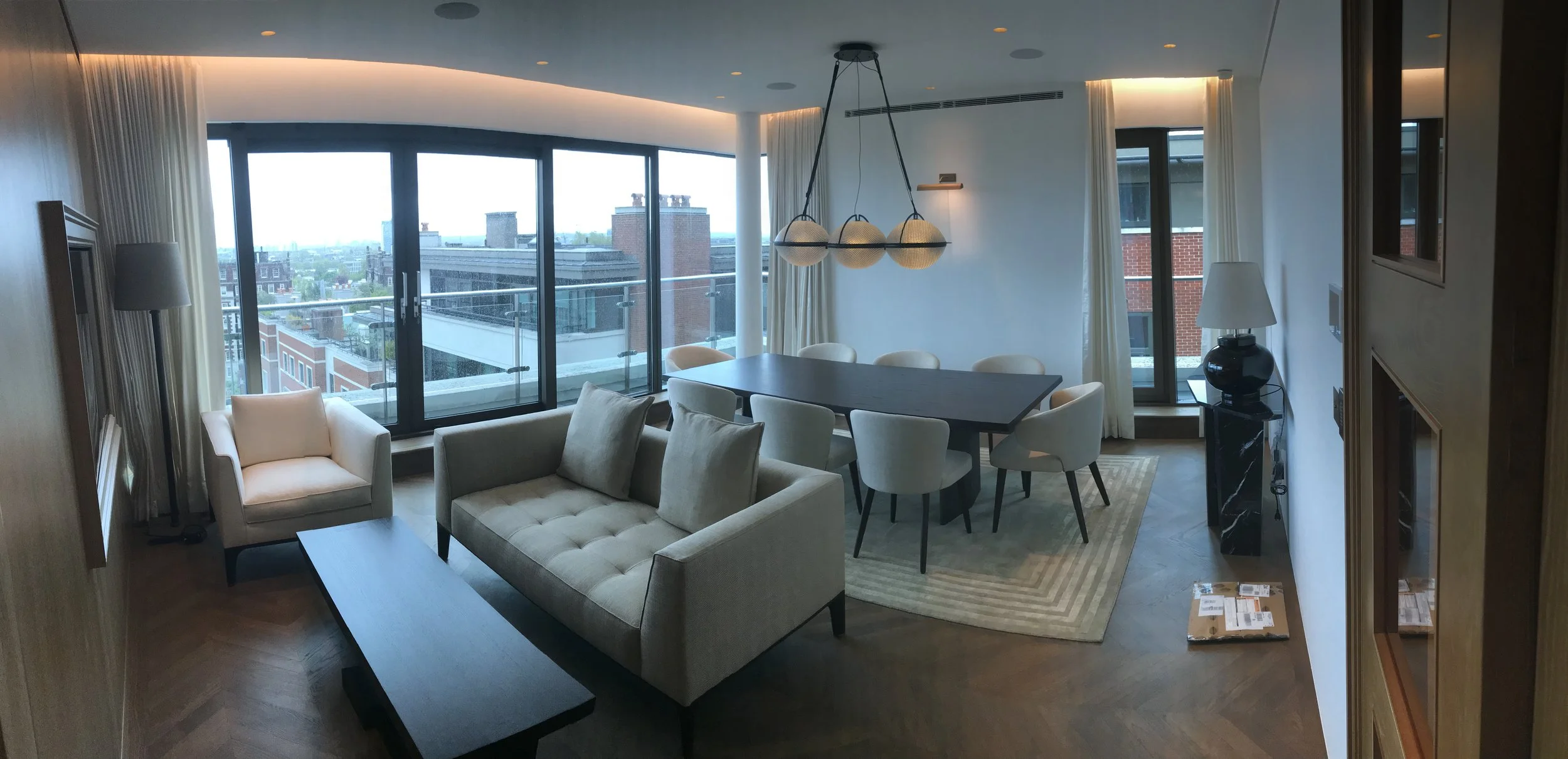 Living and dining area with large windows, beige sofa, white armchair, black dining table, cream chairs, hanging light fixture, and city view.