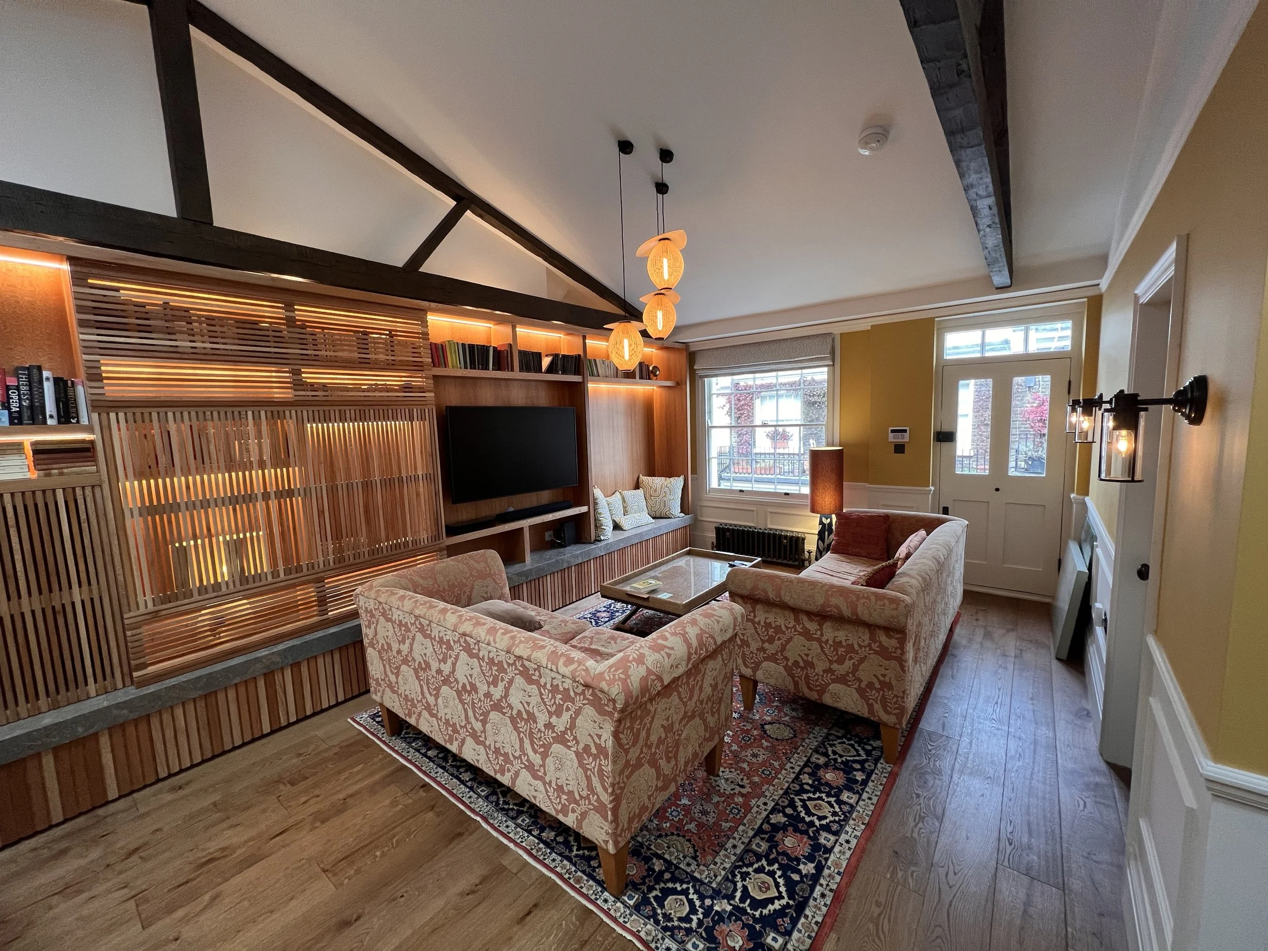 Living room with a patterned sofa, a large flat-screen TV on a wooden wall unit, pendant and wall lighting, windows, and hardwood floors.