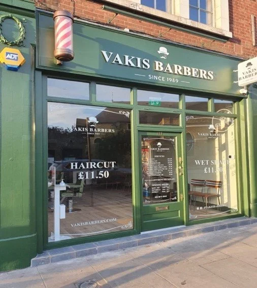 Front view of Vakis Barbershop with green facade, large glass windows, and door; signage indicating services and prices, such as haircut for £11.50 and wet shave.