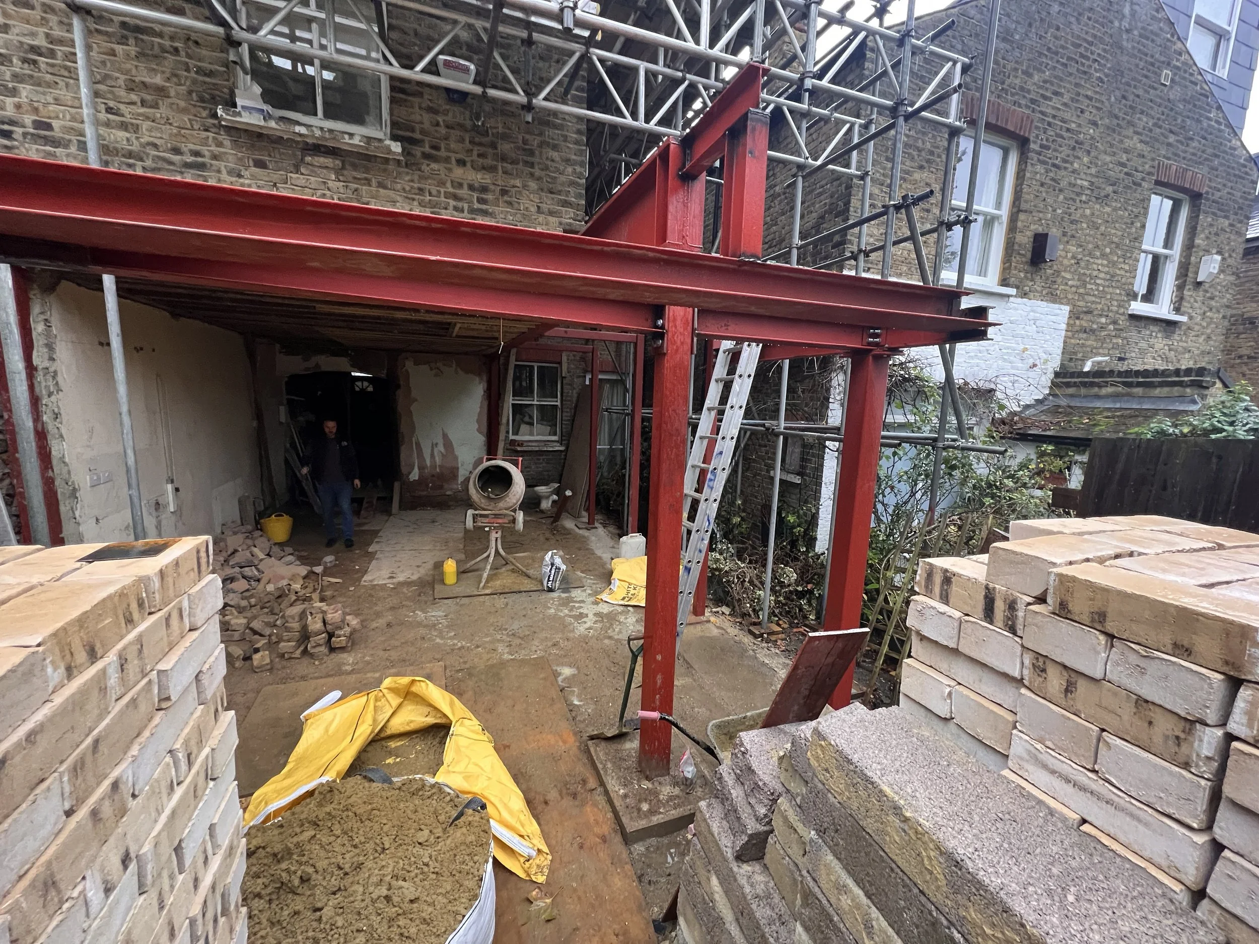Construction site with scaffolding, a worker, building materials, bricks, and a brick house in the background.