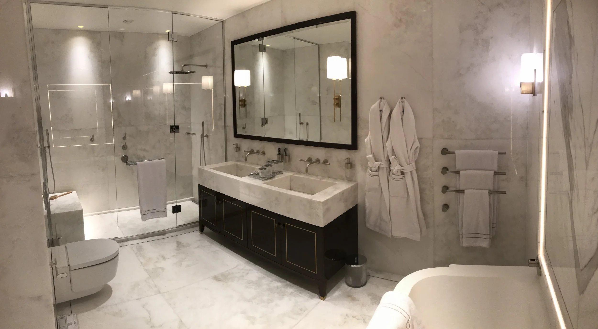 A luxurious bathroom featuring marble walls and floors, a glass-enclosed shower area with modern fixtures, a double vanity with a large mirror, wall-mounted lights, and neatly hung white towels.