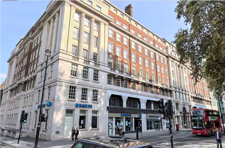 A multi-story building with a bank branch on the ground floor, situated on a city street corner with pedestrians, traffic lights, and a red double-decker bus parked nearby.