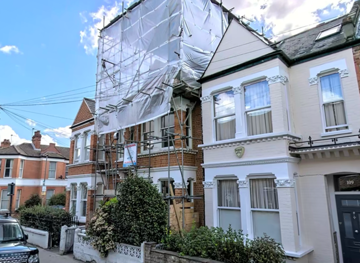A residential street scene showing a house under renovation, with scaffolding and protective white sheeting covering part of the building, next to a white Victorian-style house.