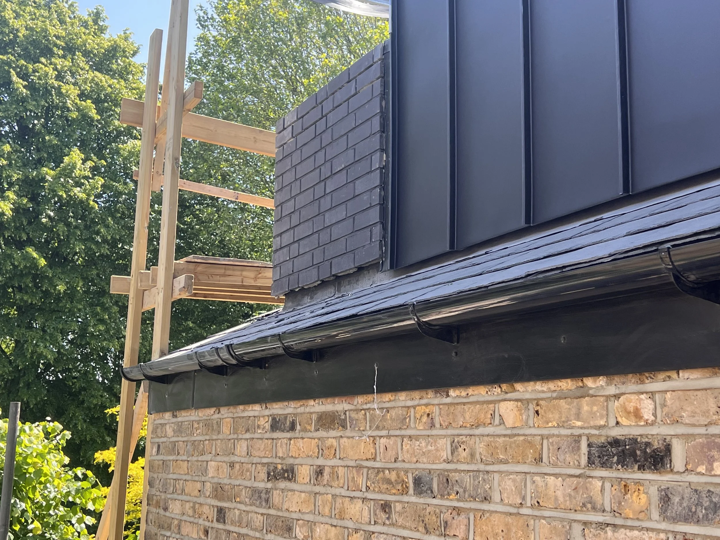 A house under construction showing gray brick chimney, black metal siding, and a black gutter system with wooden scaffolding in the background, and trees with green leaves.