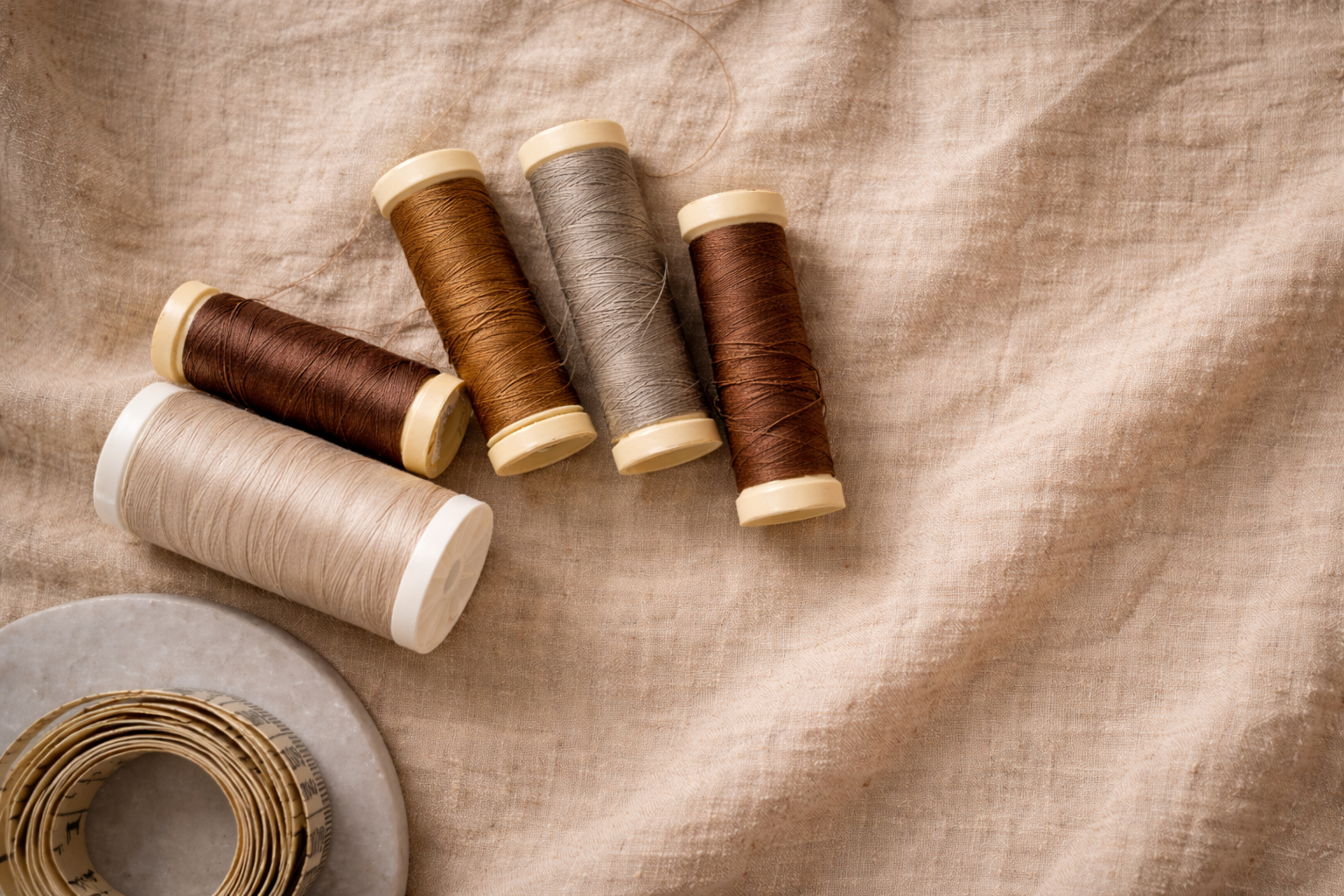 Six spools of sewing thread in various shades of beige, brown, and gray, resting on a textured beige fabric surface, with a roll of measuring tape nearby.