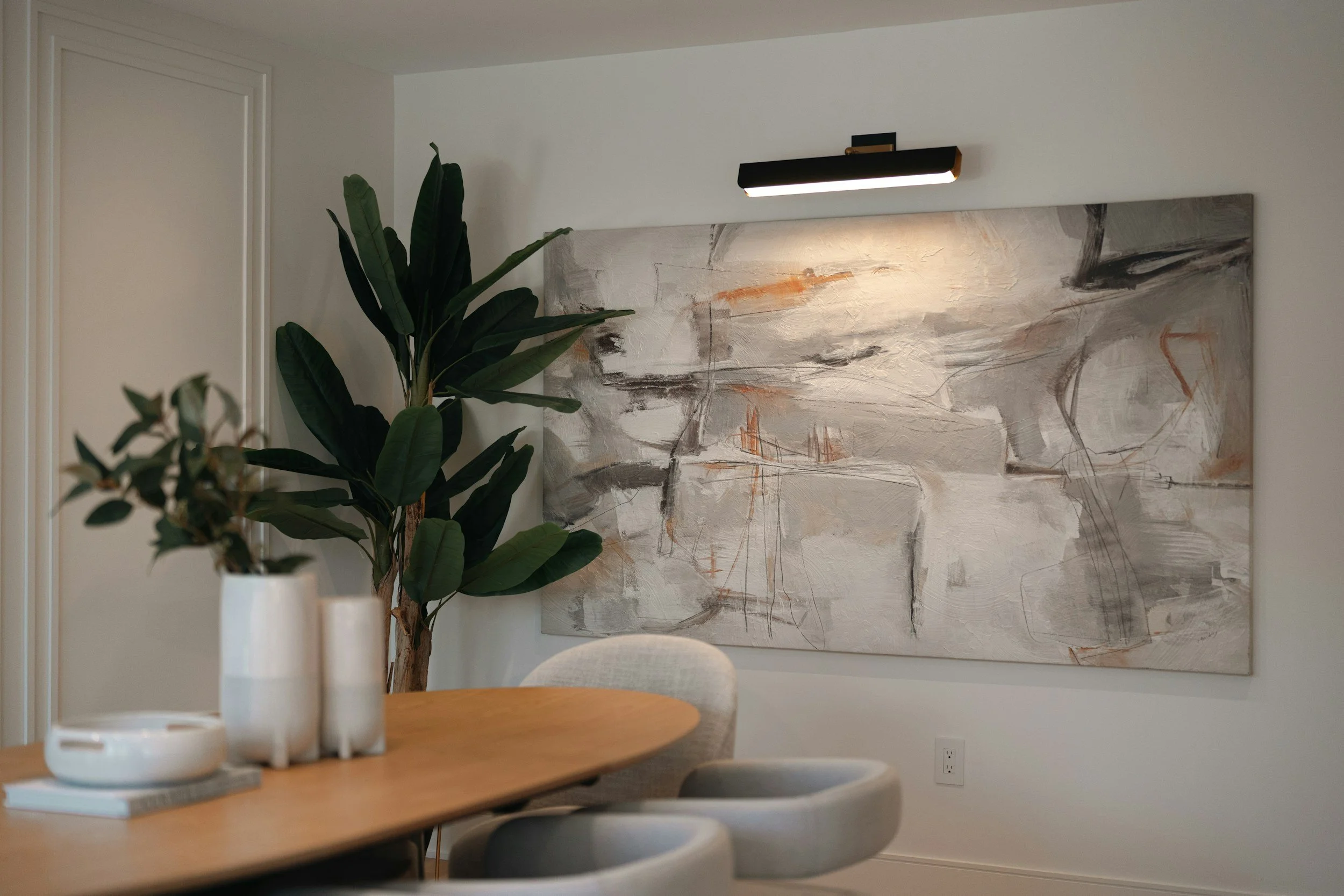 A modern dining room with a wooden table, white vases, and a large abstract painting on the wall. A tall green plant is next to the artwork, and a black wall-mounted light fixture illuminates the painting.