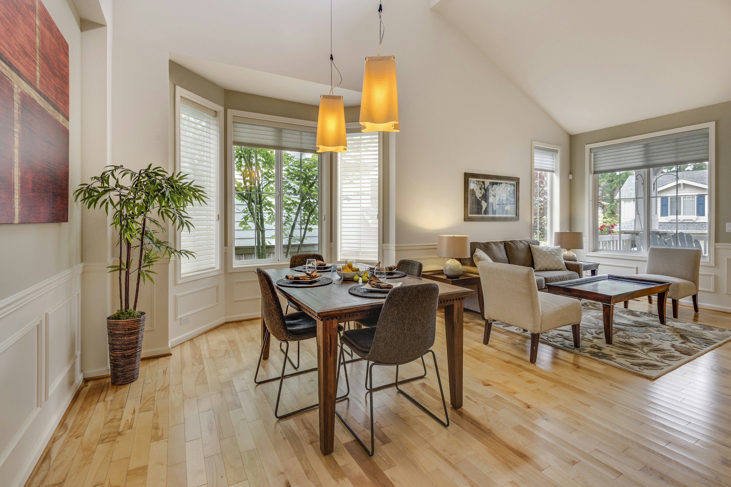 Open-concept living and dining area with large windows, hardwood floors, a dining table with chairs, a sofa, and warm lighting fixtures.