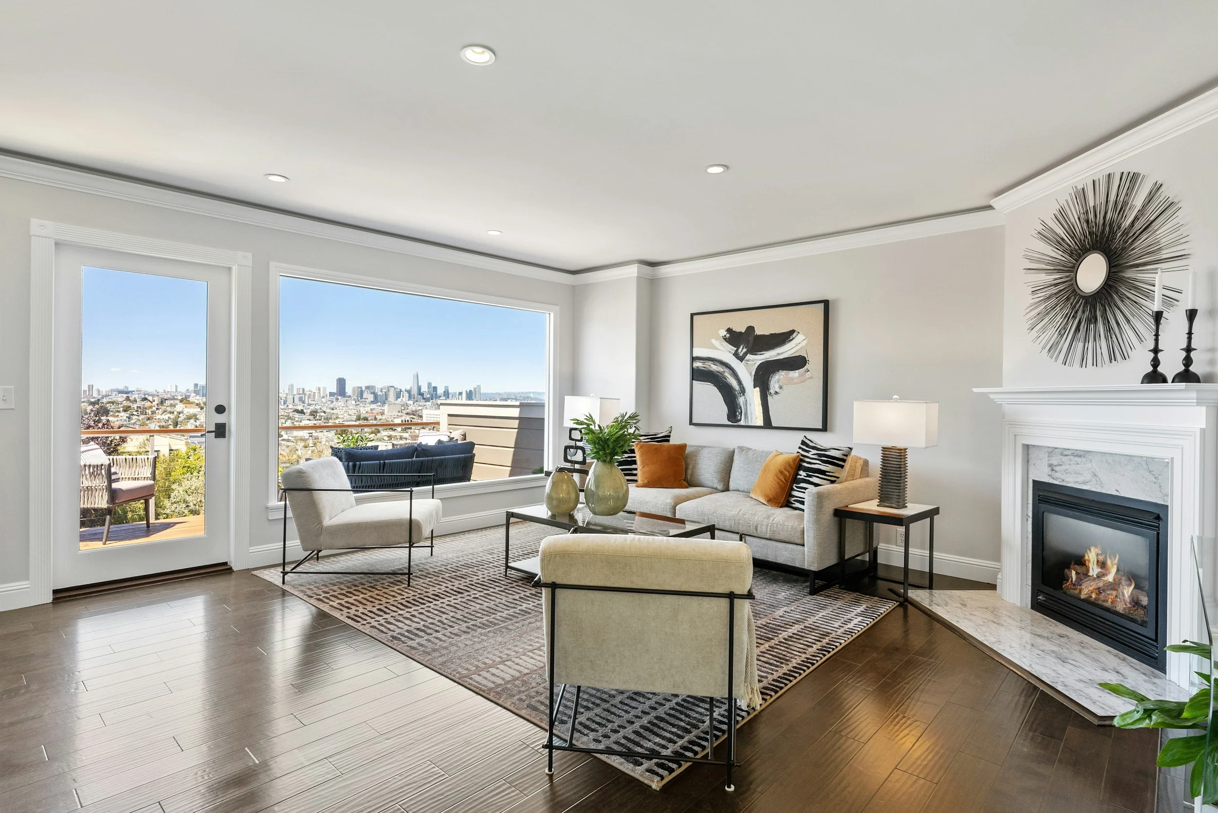 Living room with large windows showing city skyline, beige sofa with orange and black pillows, white armchair, coffee table with vases, artwork on the wall, fireplace, and ceiling lights.