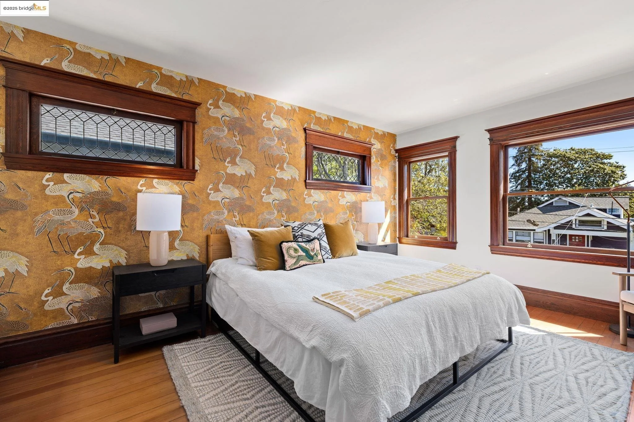 Cozy bedroom with a white bed, gold and white pillows with bird and peacock patterns, wooden window frames, a mustard yellow wallpaper with white heron patterns, and two large windows showing a view of trees and neighboring houses.