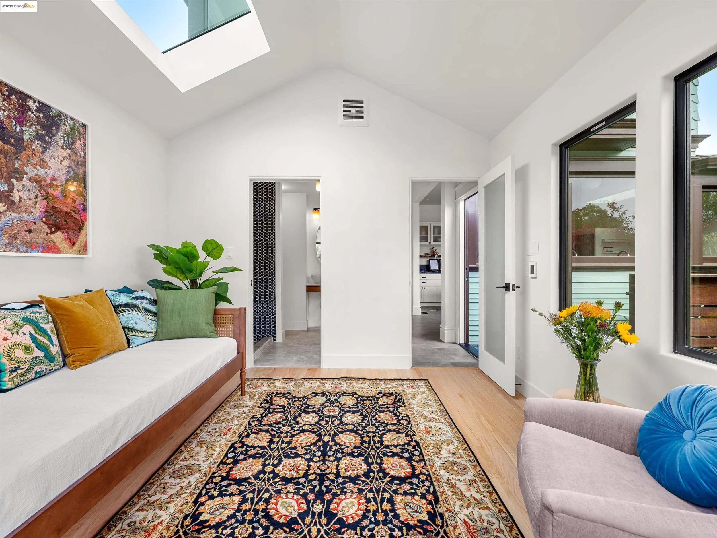 Living room with white walls and ceiling, skylight, large windows, a colorful abstract artwork on the wall, a white sofa with patterned and solid pillows, a green plant, a vase of yellow flowers, a decorative rug, and doorway leading to other rooms.