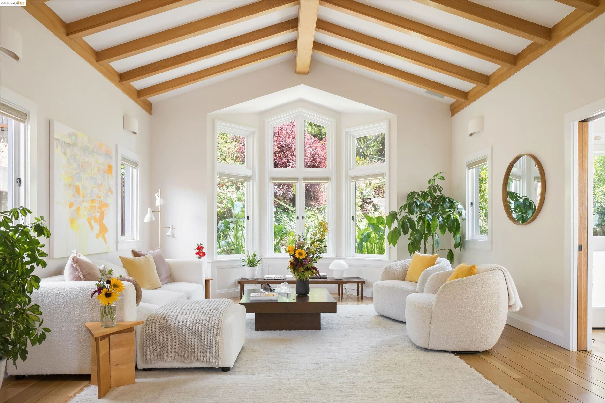 Bright living room with large bay window, white walls, and wooden vaulted ceiling with exposed beams. Contains white upholstered furniture, with yellow and beige pillows, a dark wood coffee table with a vase of colorful flowers, potted plants, wall art, round mirror, and lamps, with sunlight streaming through the windows showcasing greenery outside.