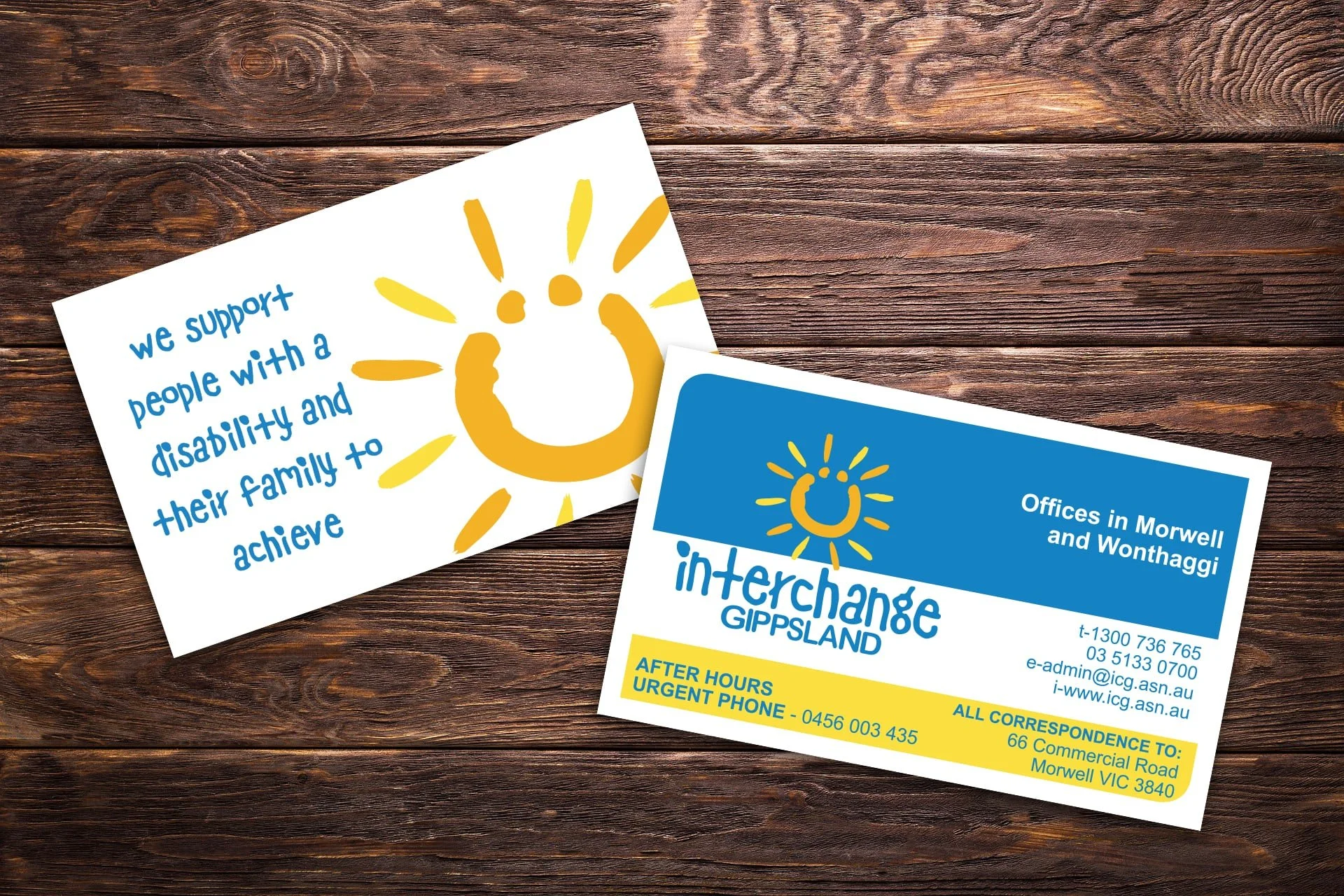 Two business cards for Interchange Gippsland, with a yellow sun and blue text, placed on a wooden surface.