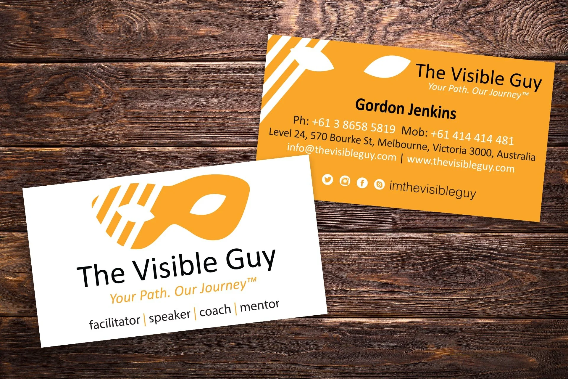 Two business cards for The Visible Guy, one with a white background and one with an orange background. The white card features a stylized orange and white logo of a mask and the company name in black, along with some tagline text and titles. The oran