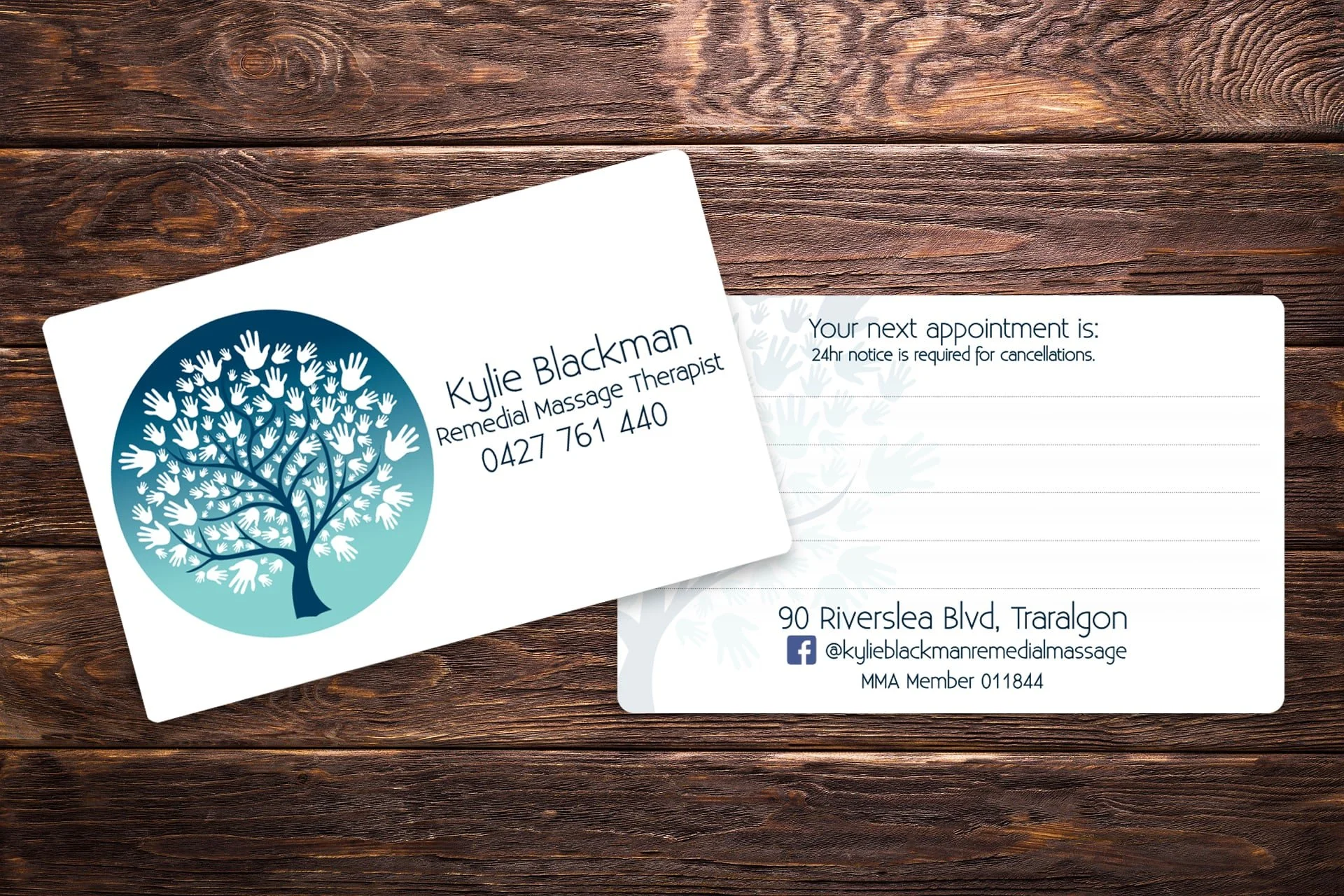 Business card for Kylie Blackman, Remedial Massage Therapist, featuring a blue gradient tree logo with white handprint leaves, contact number, address, Facebook handle, and membership number on a wooden surface background.