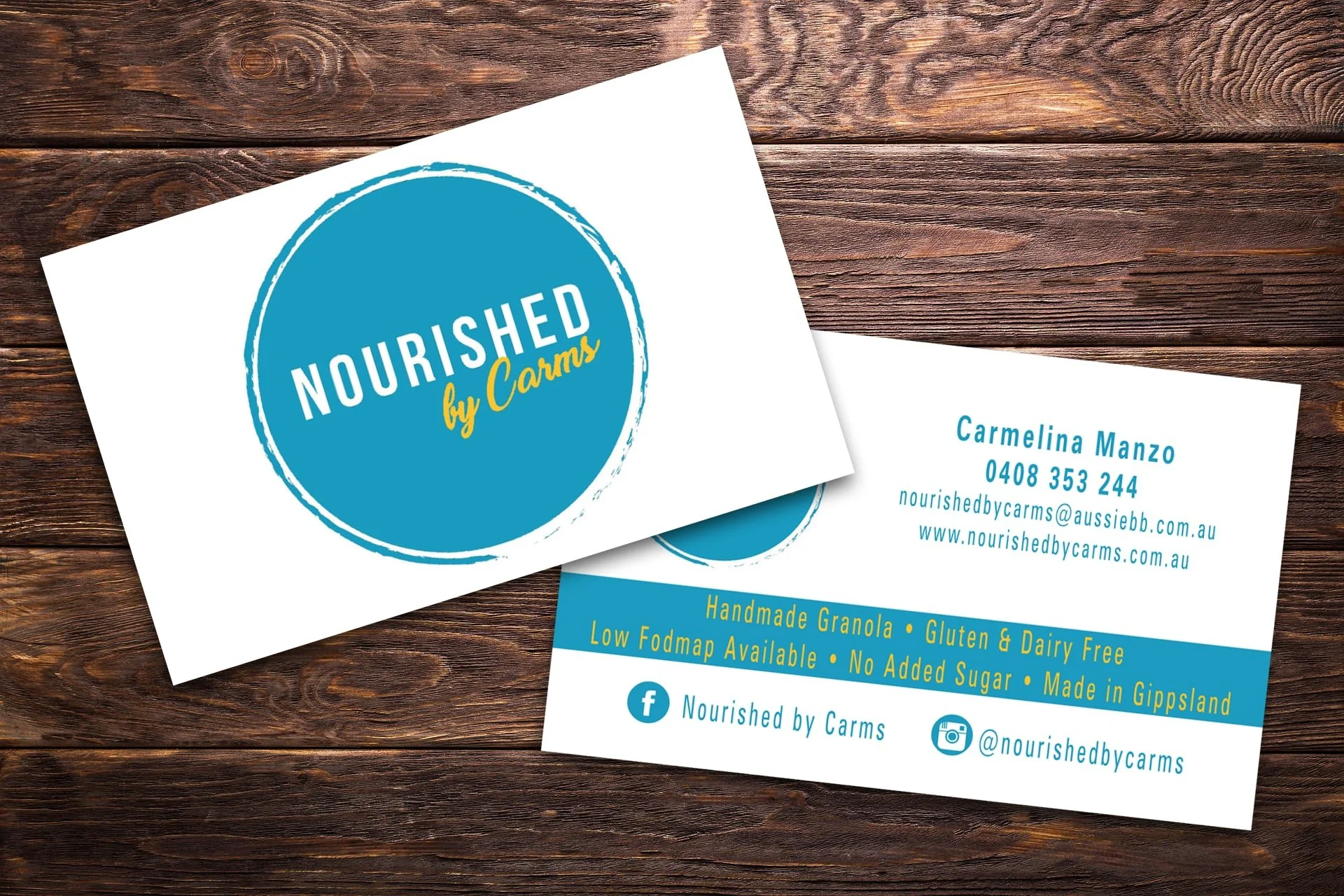 Business cards for Nourished by Carms placed on a wooden surface. One card displays the company logo, a blue circle with white and yellow text, and the other card provides contact details and product information.