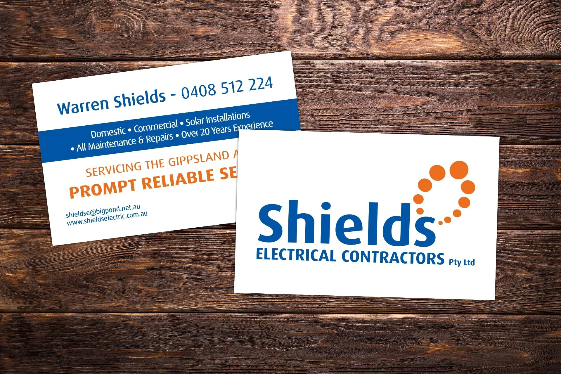 Business cards for Shields Electrical Contractors Pty Ltd, placed on a wooden surface. One card displays the company's logo with a stylized orange dotted circle and the name in blue text, while the other card contains contact information, services of
