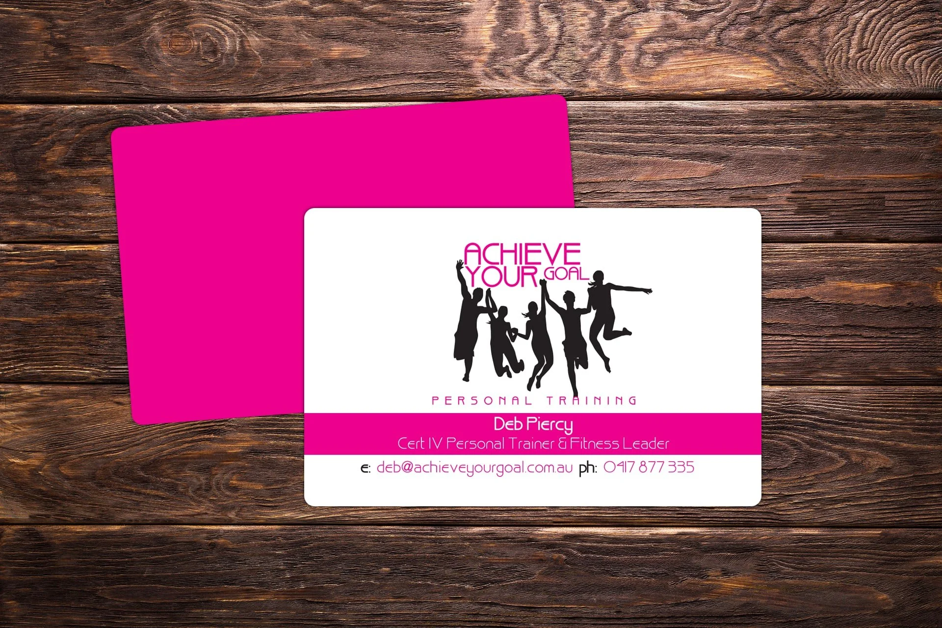 Business card for Deb Piercy, a personal trainer and fitness leader, with pink and white design, silhouetted people jumping, and contact information, placed on a wooden surface.