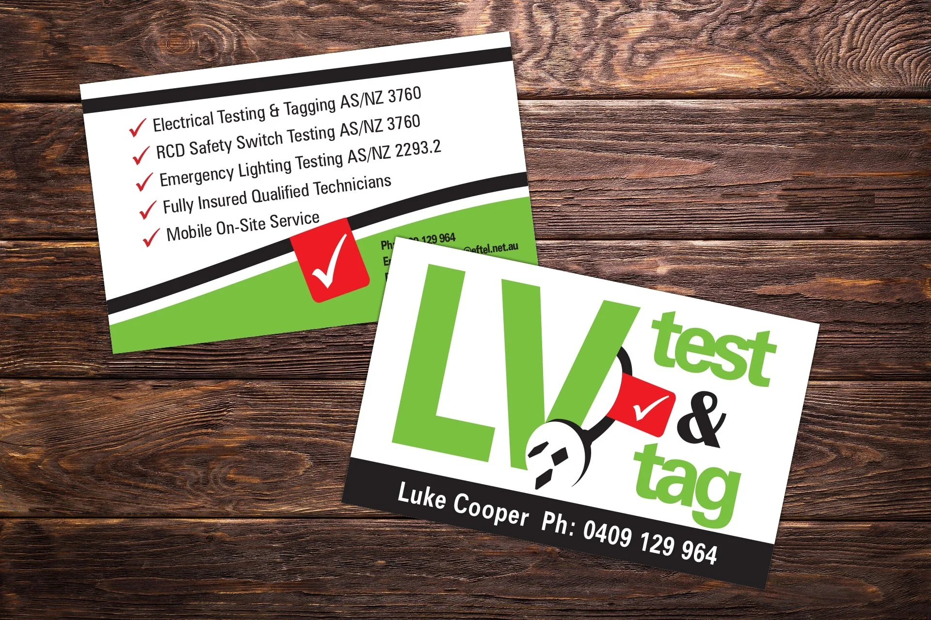 Two printed cards on a wooden surface, one partially overlapping the other. The top card lists services such as electrical testing, switch testing, emergency lighting testing, and on-site service, with a red checkmark icon next to each. The bottom ca
