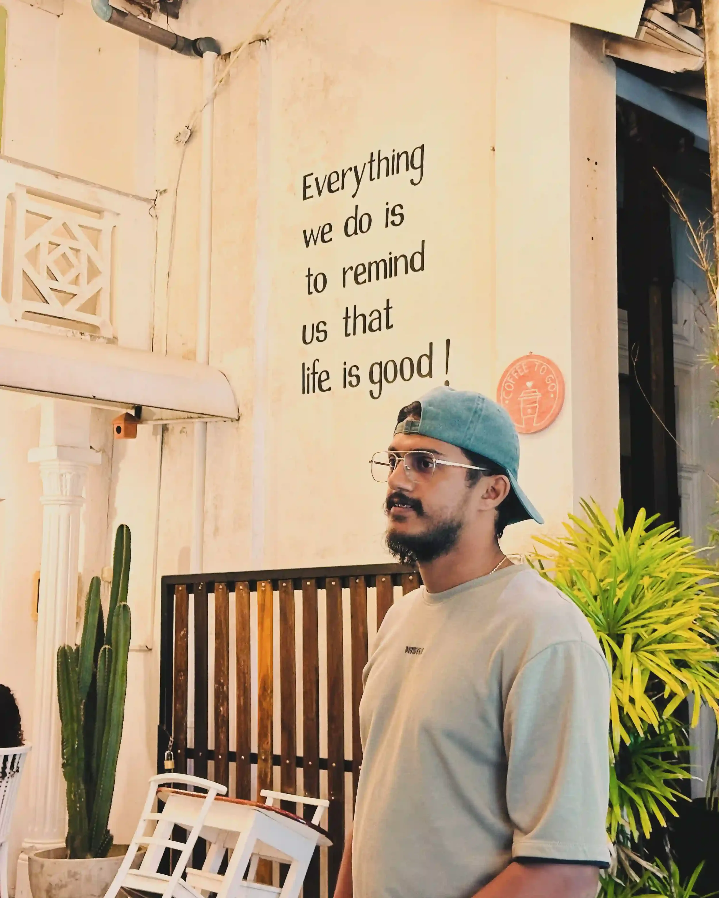 A young man wearing glasses and a backwards baseball cap standing outdoors at night, with plants and a white table nearby. There is a wall with a quote that reads, 'Everything we do is to remind us that life is good!'