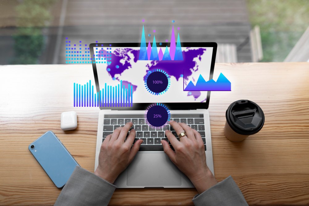 Person using a laptop with digital data graphs and world map overlay, a smartphone, wireless earbuds, and a takeaway coffee cup on a wooden desk.