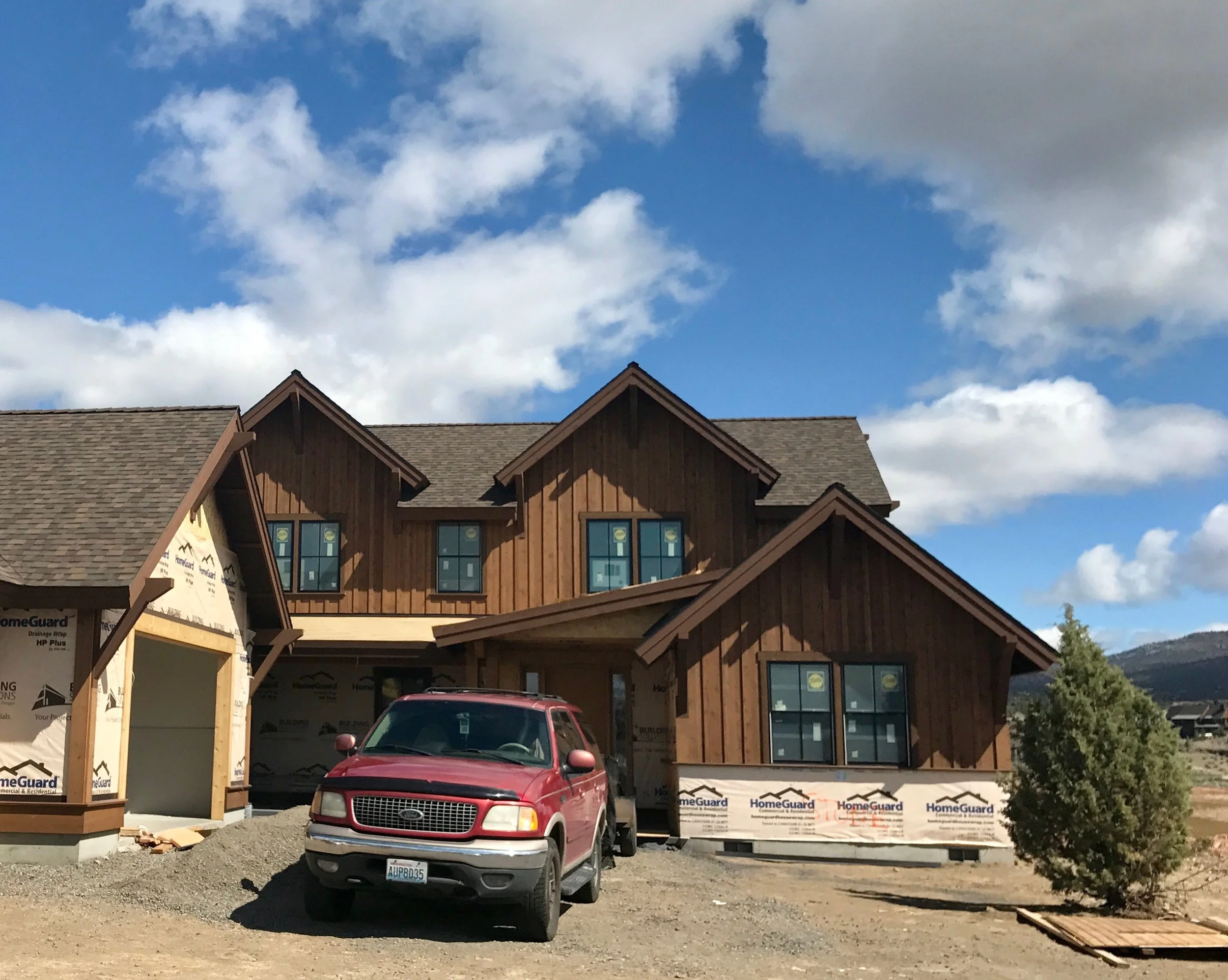 A house under construction with a red SUV parked in front. The house has wooden siding and multiple windows, some with stickers. The sky is partly cloudy with blue sky visible in the background.