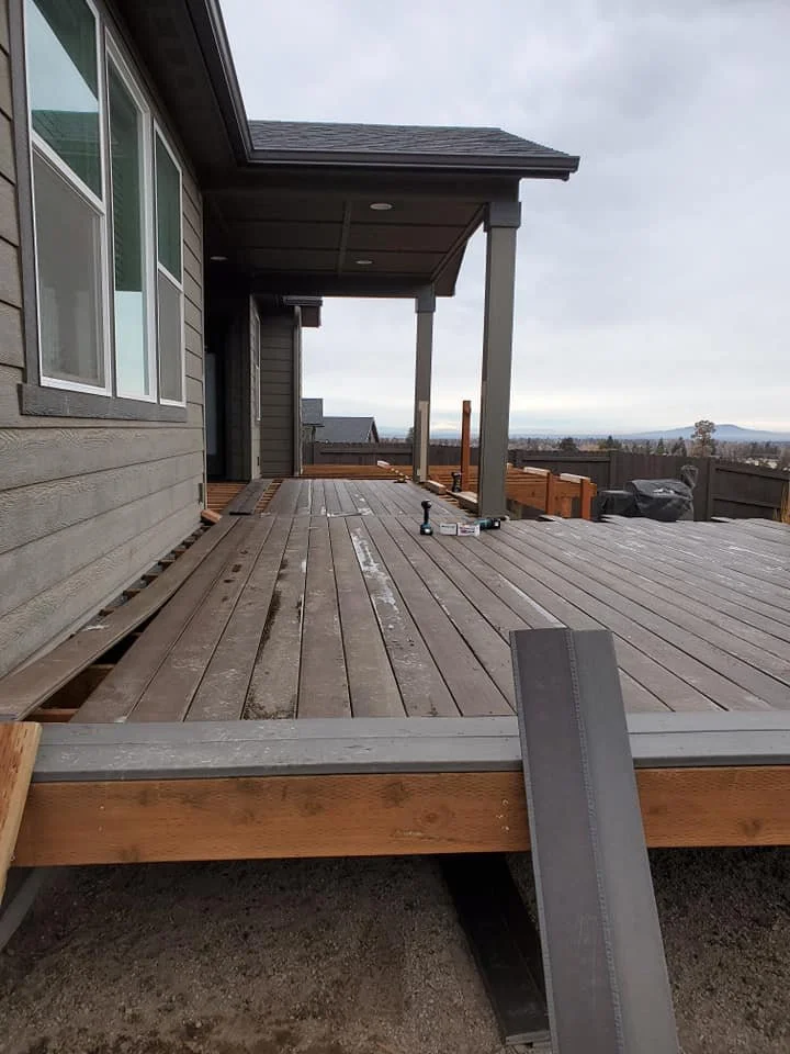 View of a partially constructed wooden deck attached to a house with gray siding, showing tools and some wooden rails, under a cloudy sky.