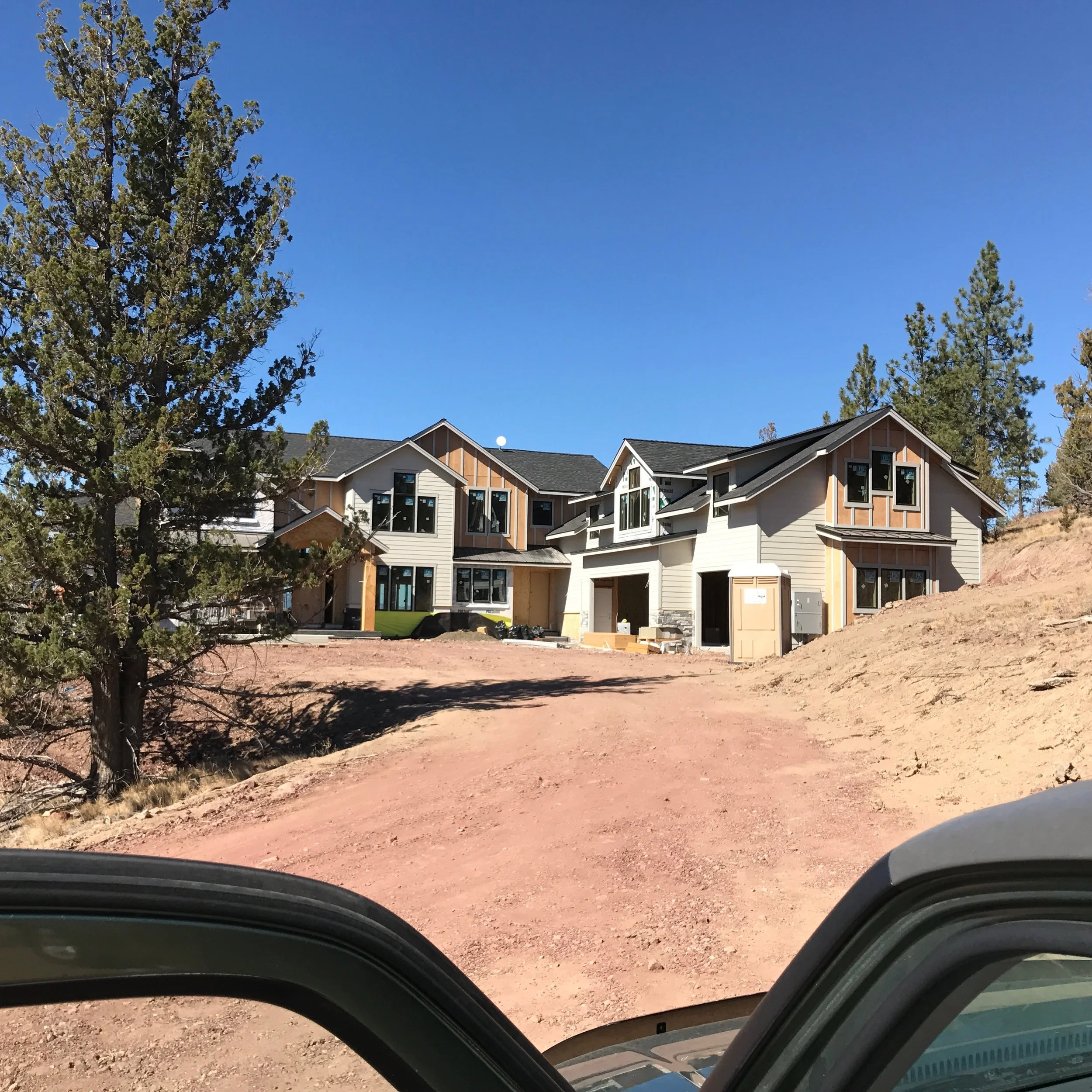 Newly constructed houses on a dirt lot under a clear blue sky with some trees around.