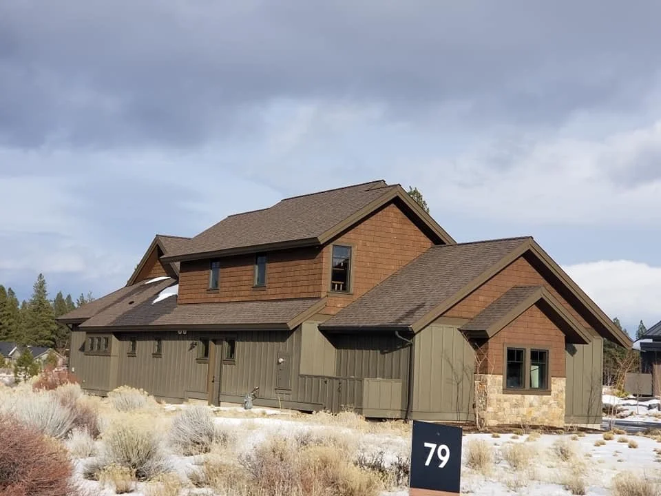 A two-story house with brown and green exterior, surrounded by a desert landscape with dry bushes and snow, under a cloudy sky.