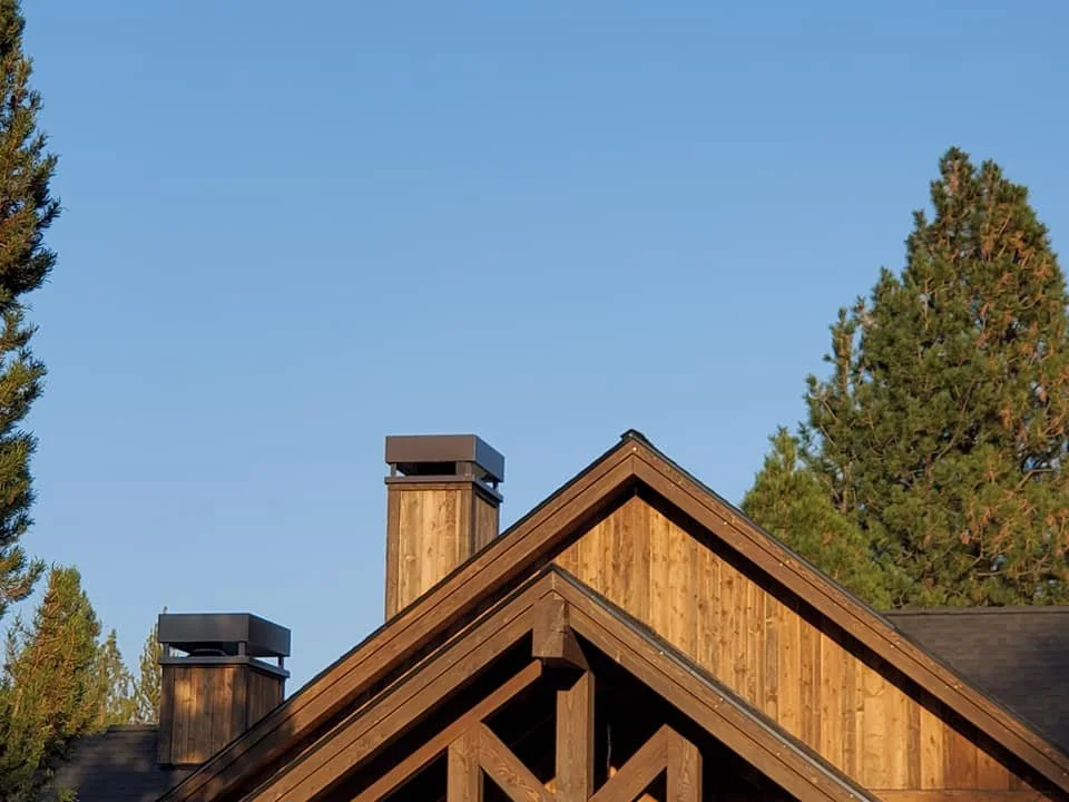 Close-up of a wooden house roof with two chimney stacks and evergreen trees in the background under a clear blue sky.