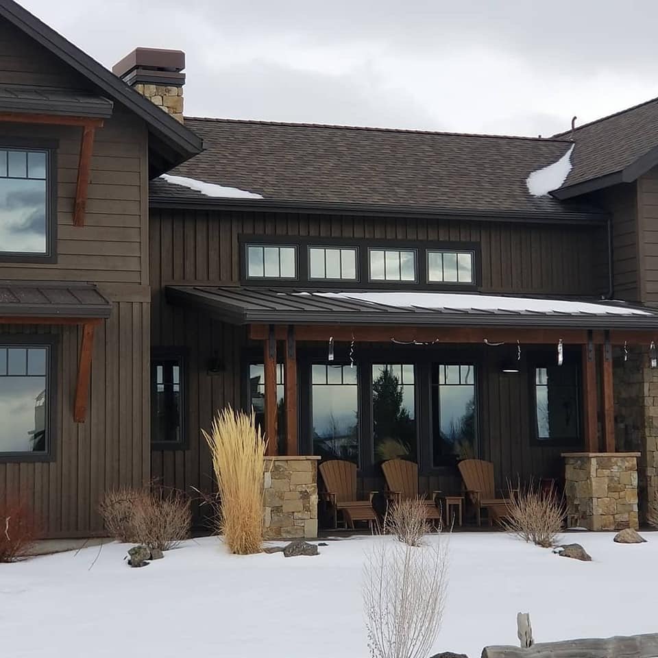 A house with a wooden exterior, stone accents, and black-framed windows, with snow on the ground and a cloudy sky.