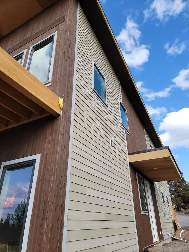 A modern, multi-story house with wood and beige siding, featuring multiple windows and two small overhangs on the second floor, against a background of a partly cloudy blue sky.