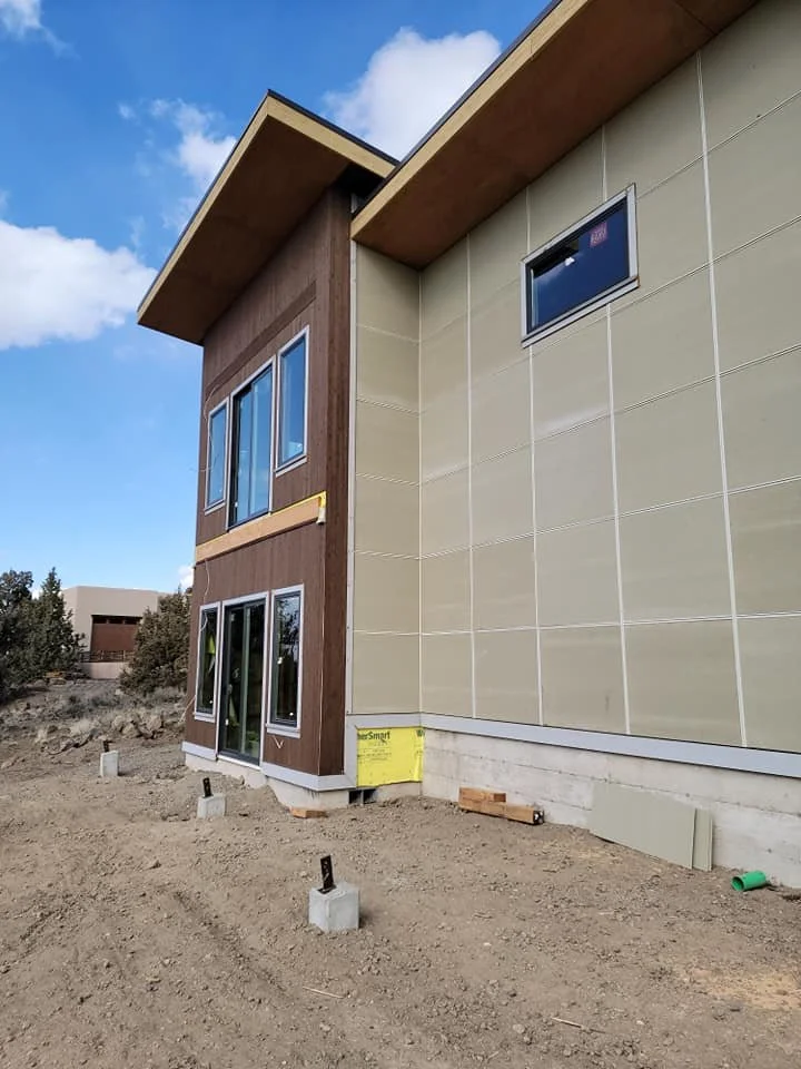 Side view of a modern house under construction with beige siding, wooden accents, a small window, and a sliding glass door. The area around the house is bare dirt with construction supports in place.