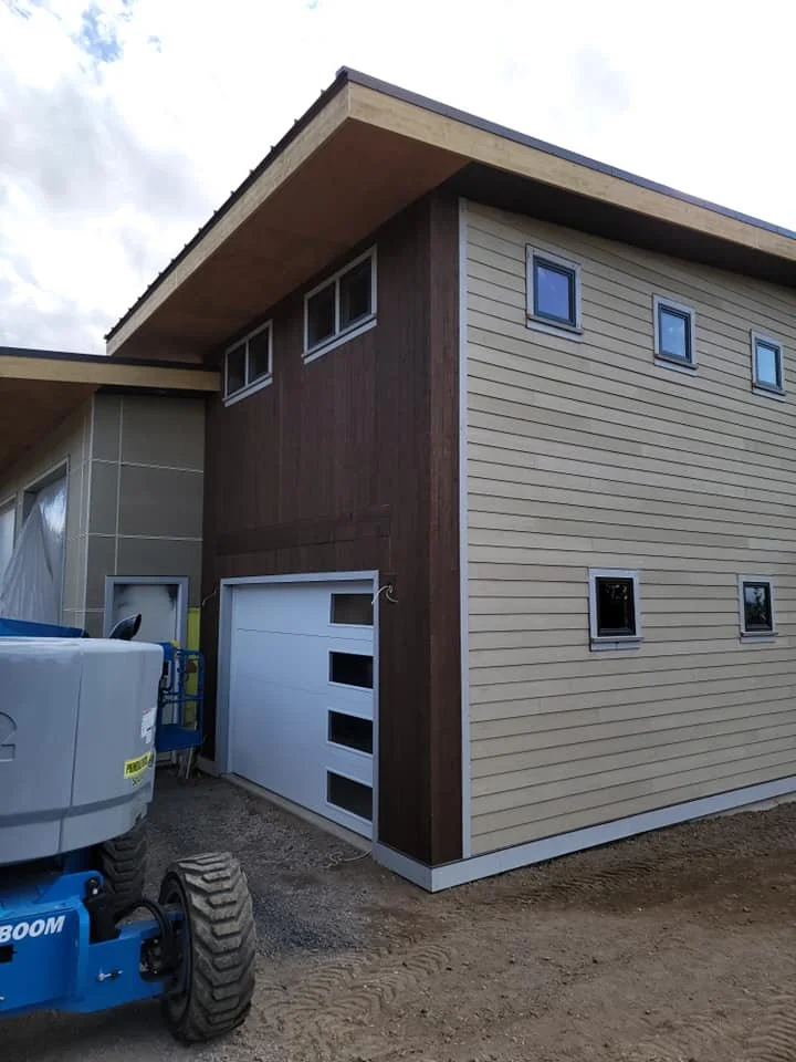 A modern two-story house under construction with beige and brown exterior siding, multiple small windows, and a white garage door, on a dirt lot.