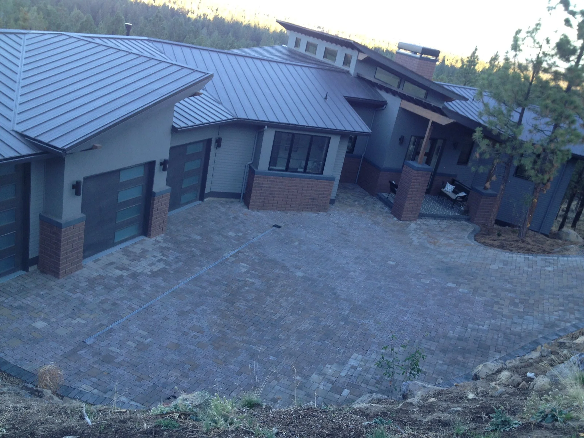 Modern house with a metal roof, brick accents, a paved driveway, and a small porch area with seating, surrounded by trees.