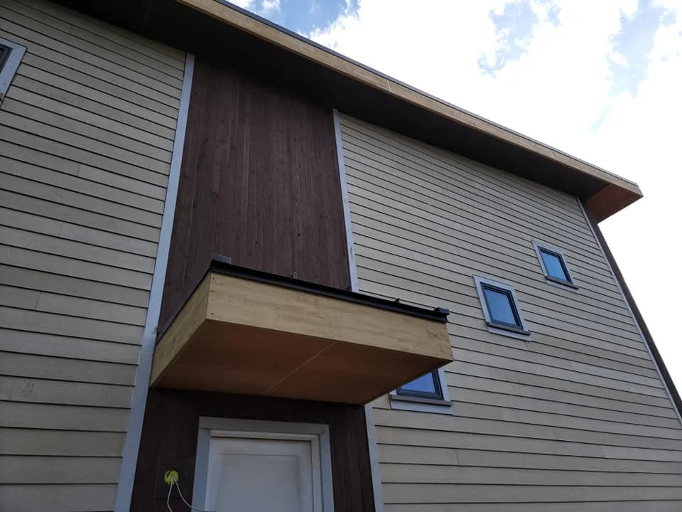 Exterior view of a modern house with beige and brown siding, featuring a small balcony and multiple small windows.