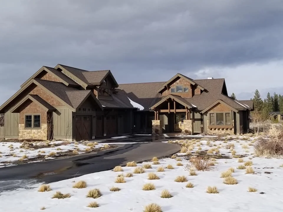 Large house with a stone and wooden exterior, surrounded by a snow-covered lawn with patches of dry grass, under a cloudy sky.