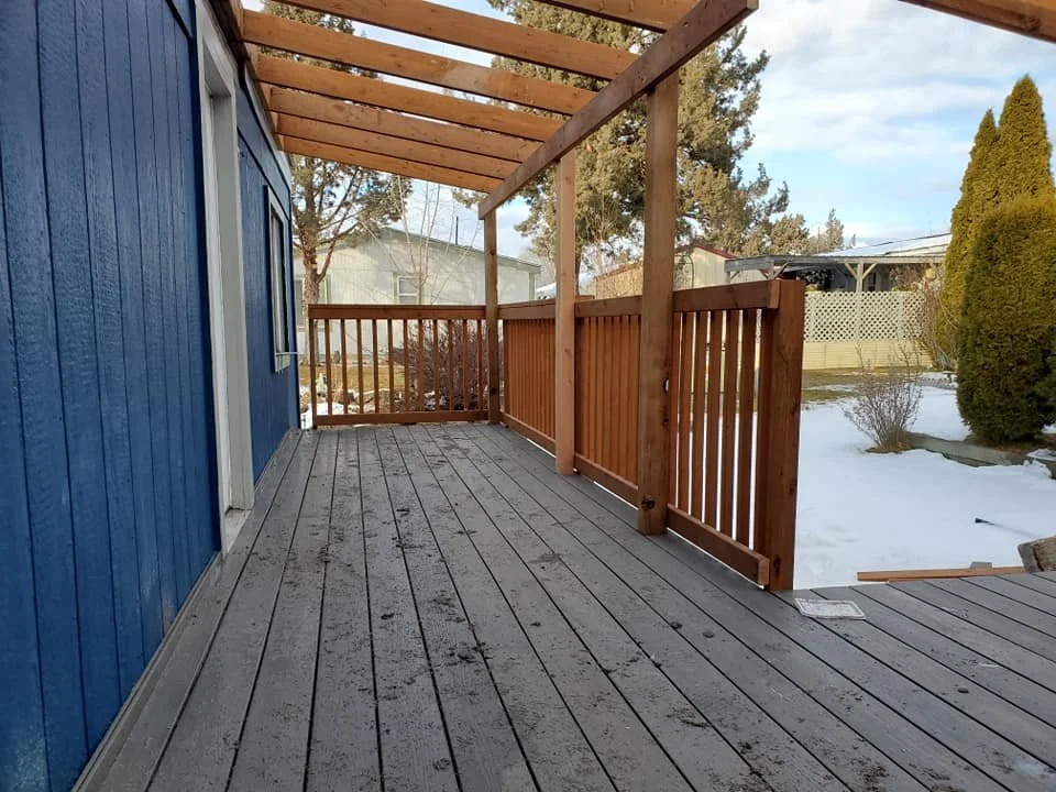 Wooden deck with a partial roof under construction, attached to a blue house, overlooking a snow-covered yard with trees and shrubs.