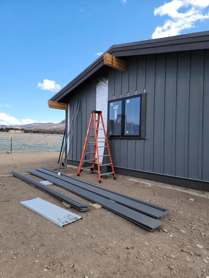 Construction site with a dark gray house, orange ladder, and building materials on the ground outside, under a partly cloudy sky.