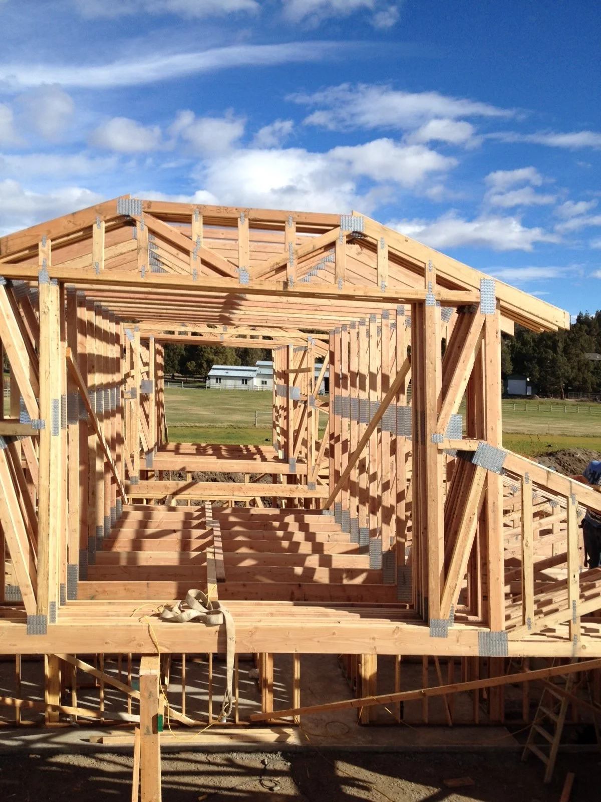 Wooden framing structure of a house under construction, with multiple rectangular openings, set against a bright blue sky with clouds and a rural landscape with trees and buildings in the background.