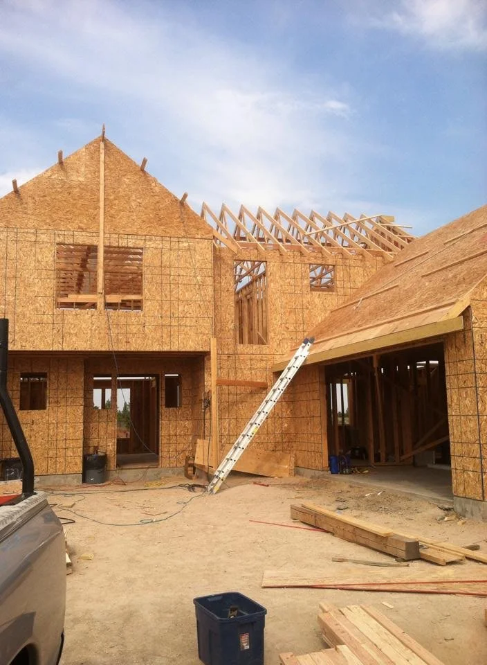 Construction site of a house with wooden framing and plywood sheathing, partially built roof with trusses, ladder leaning against the structure, and construction materials on the ground.