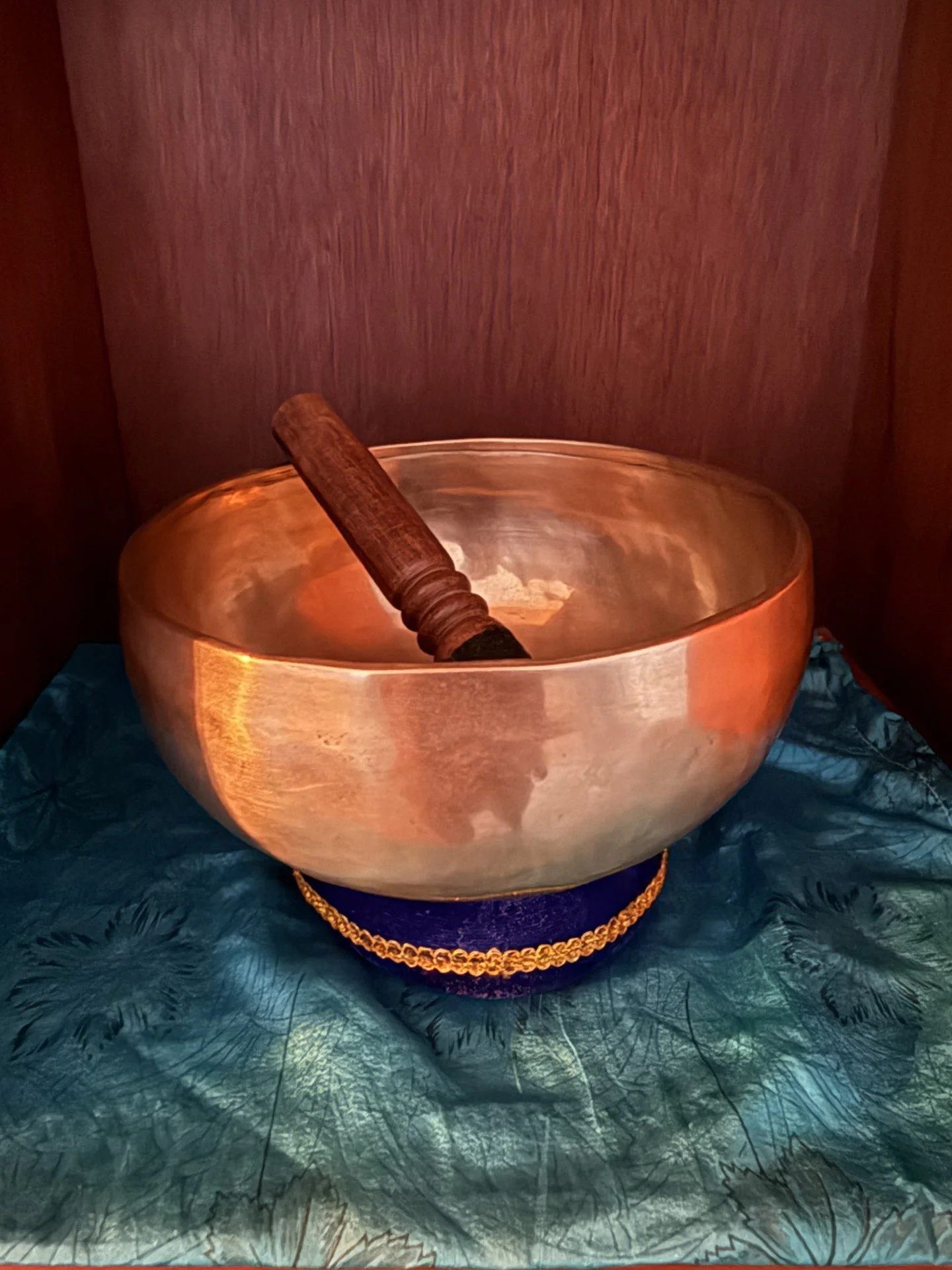 A large singing bowl made of copper or brass, with a wooden mallet resting inside, placed on a decorative stand with a gold-colored chain around its base, set on a textured teal fabric.